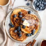 Overhead view of a plate of three pancakes topped with butter, maple syrup, and blueberries, next to a bowl of blueberries, cinnamon sticks, and a cup of coffee