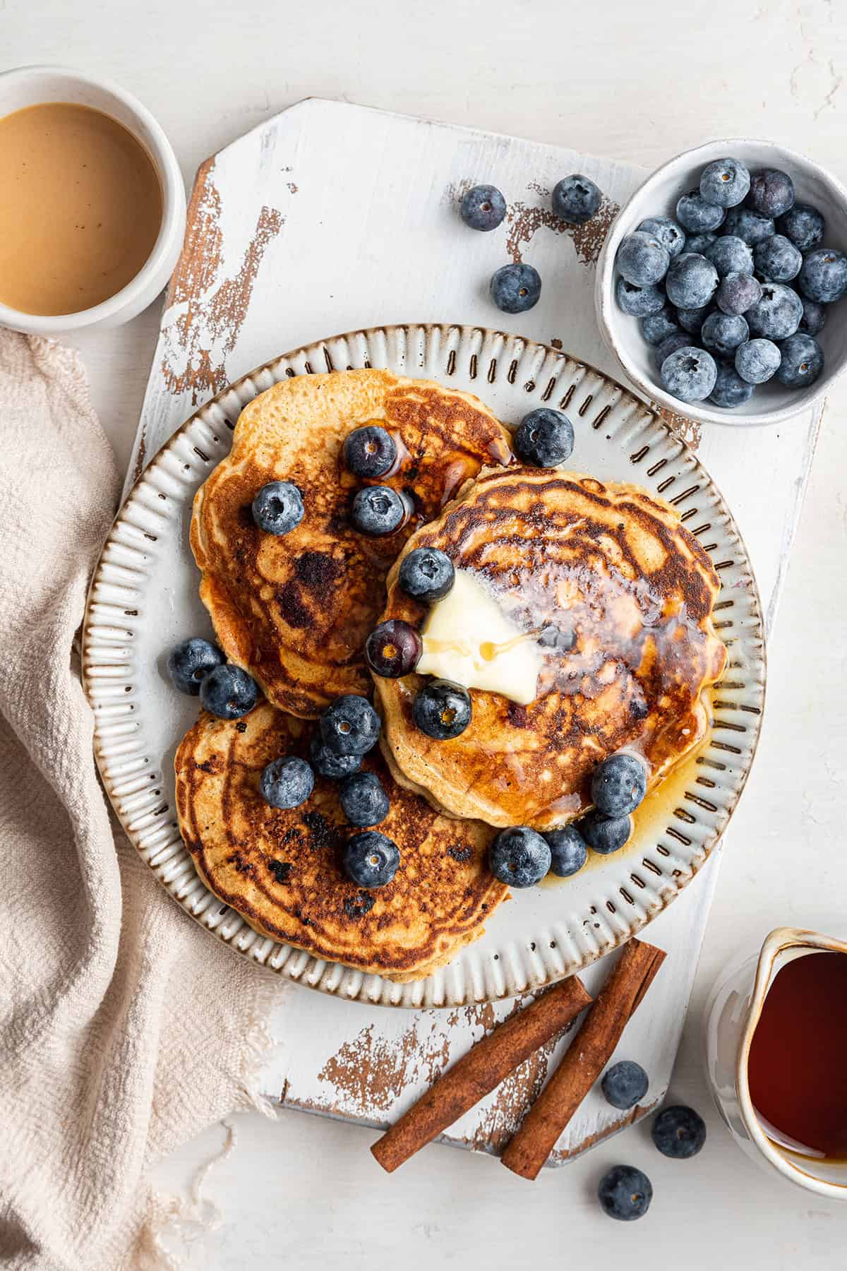 These perfect gluten-free blueberry pancakes are light, fluffy, tart, and sweet, and only take 15 minutes to make! Overhead view of a plate of three pancakes topped with butter, maple syrup, and blueberries, next to a bowl of blueberries, cinnamon sticks, and a cup of coffee