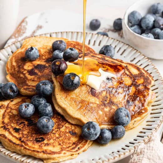 Maple syrup being poured onto a plate of three pancakes topped with blueberries and butter, with blueberries behind them