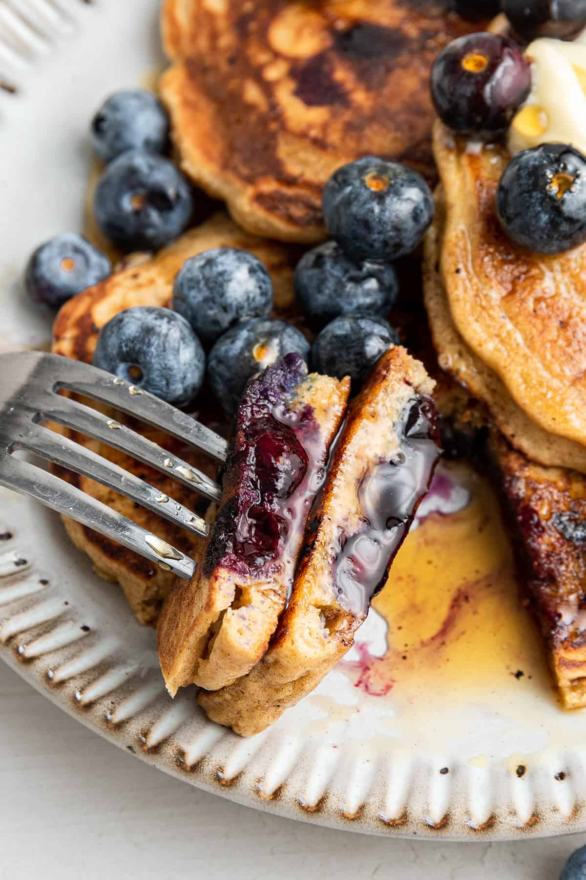 These perfect gluten-free blueberry pancakes are light, fluffy, tart, and sweet, and only take 15 minutes to make! Close up of a fork with a bite of blueberry pancakes on it, with blueberries and pancakes in the background