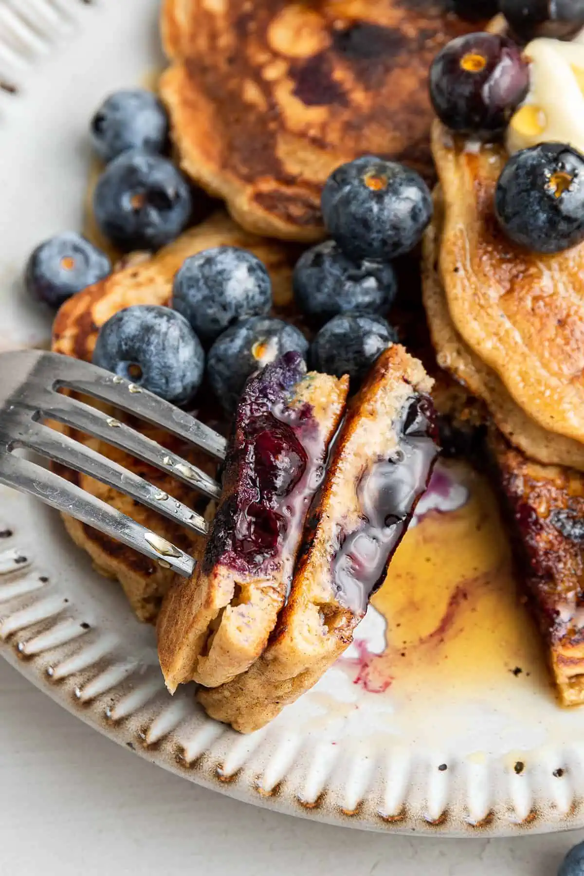 Close up of a fork with a bite of blueberry pancakes on it, with blueberries and pancakes in the background