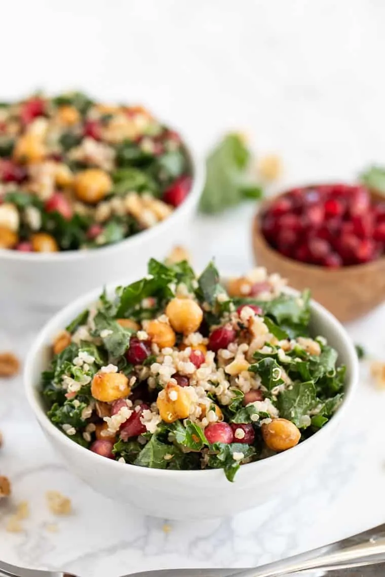 This kale pomegranate salad is a quick and easy winter salad recipe with hearty greens, quinoa, walnuts, chickpeas, and pomegranate seeds. Two bowls of kale pomegranate salad next to a smaller bowl of pomegranate seeds.