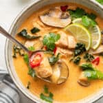 A bowl of Thai coconut curry soup, garnished with chilis, lime slices, and cilantro, with a spoon taking a bite out, and a lime in the background