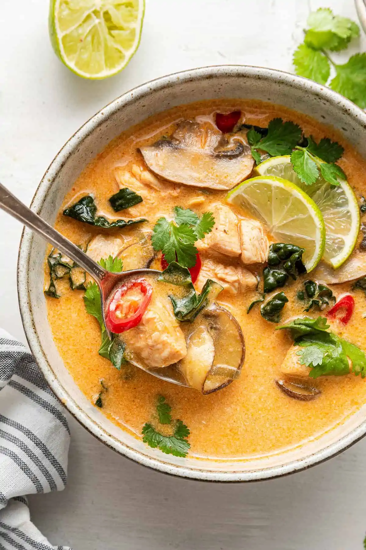 A bowl of Thai coconut curry soup, garnished with chilis, lime slices, and cilantro, with a spoon taking a bite out, and a lime in the background