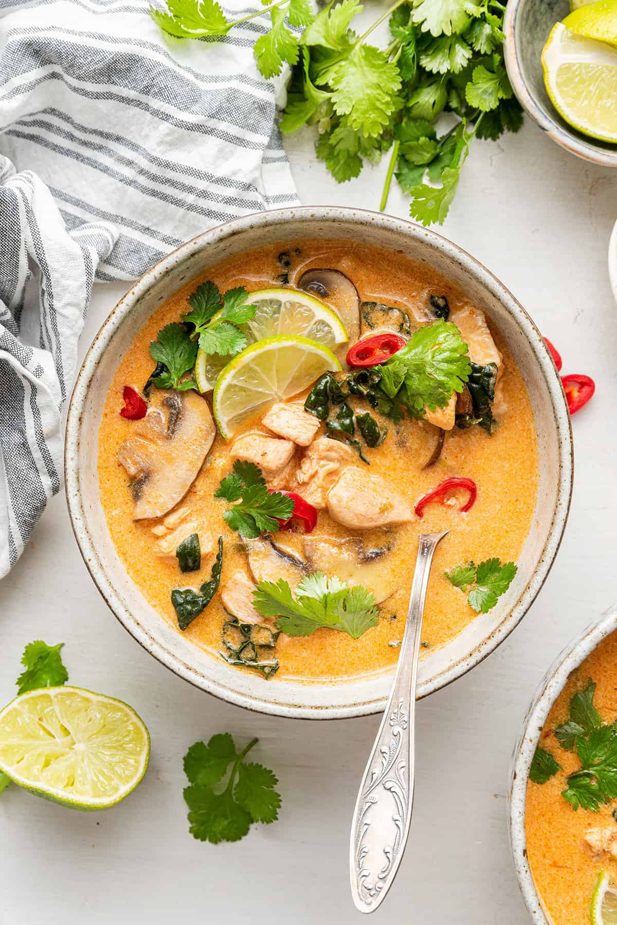 A bowl of Thai coconut curry soup, garnished with lime slices, cilantro, and chilis, with a spoon in it, next to some lime slices, cilantro, and a kitchen towel