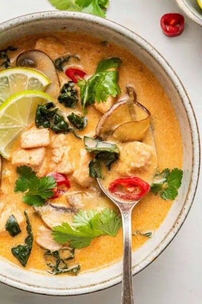 Close up of a spoon in a bowl of Thai coconut curry soup, garnished with chilis, lime slices, and cilantro