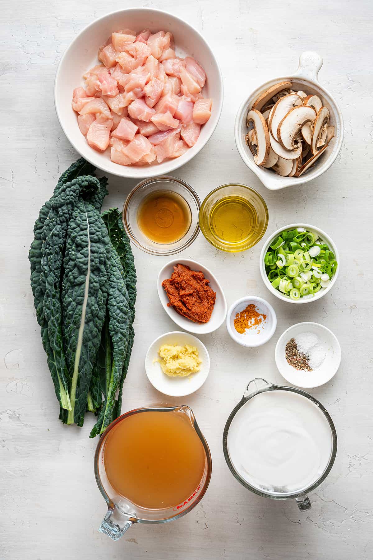 Overhead view of the ingredients needed for Thai coconut curry soup: A bowl of cubed chicken breast, a pyrex of chicken broth, a pyrex of coconut milk, a bowl of mushrooms, a bowl of green onions, a bowl of ginger, a bowl of curry paste, a bowl of oil, a bowl of fish sauce, and some kale leaves.