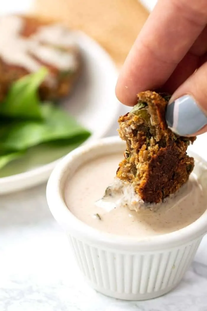 A hand dipping a lentil patty into a ramekin of dipping sauce, with more patties on a plate in the background.