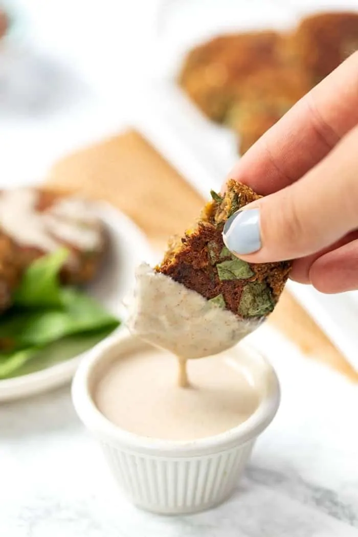 A hand dipping a lentil patty into a ramekin of dipping sauce, with more patties on a plate in the background.