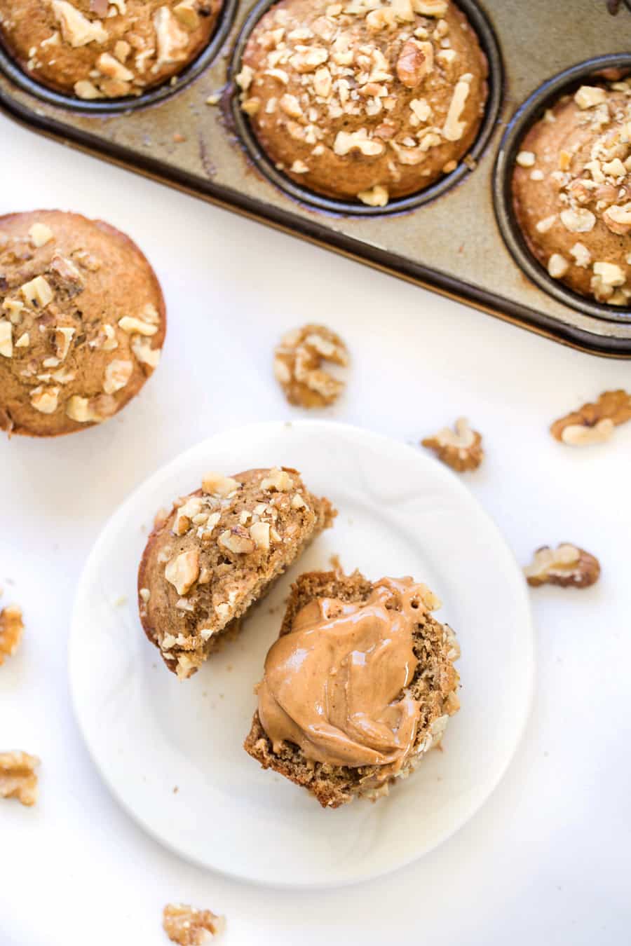 Overhead view of a banana muffin on a plate, cut in half, and topped with peanut butter, surrounded by more muffins and walnuts. 