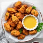 Overhead view of a serving tray filled with popcorn chicken that is garnished with parsley, with a bowl of dipping sauce on the tray, and a kitchen towel next to it