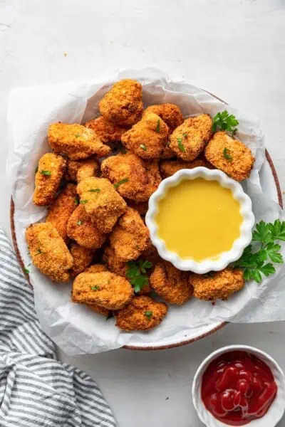 Overhead view of a serving tray filled with popcorn chicken that is garnished with parsley, with a bowl of dipping sauce on the tray, and a kitchen towel next to it