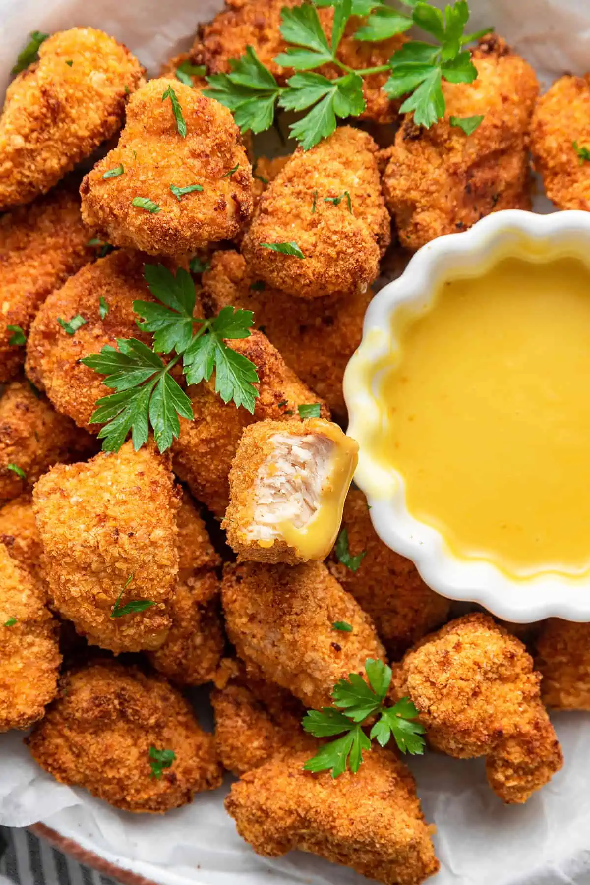 Close up of a pile of popcorn chicken garnished with parsley, with a bite taken out of one piece, and a bowl of dipping sauce