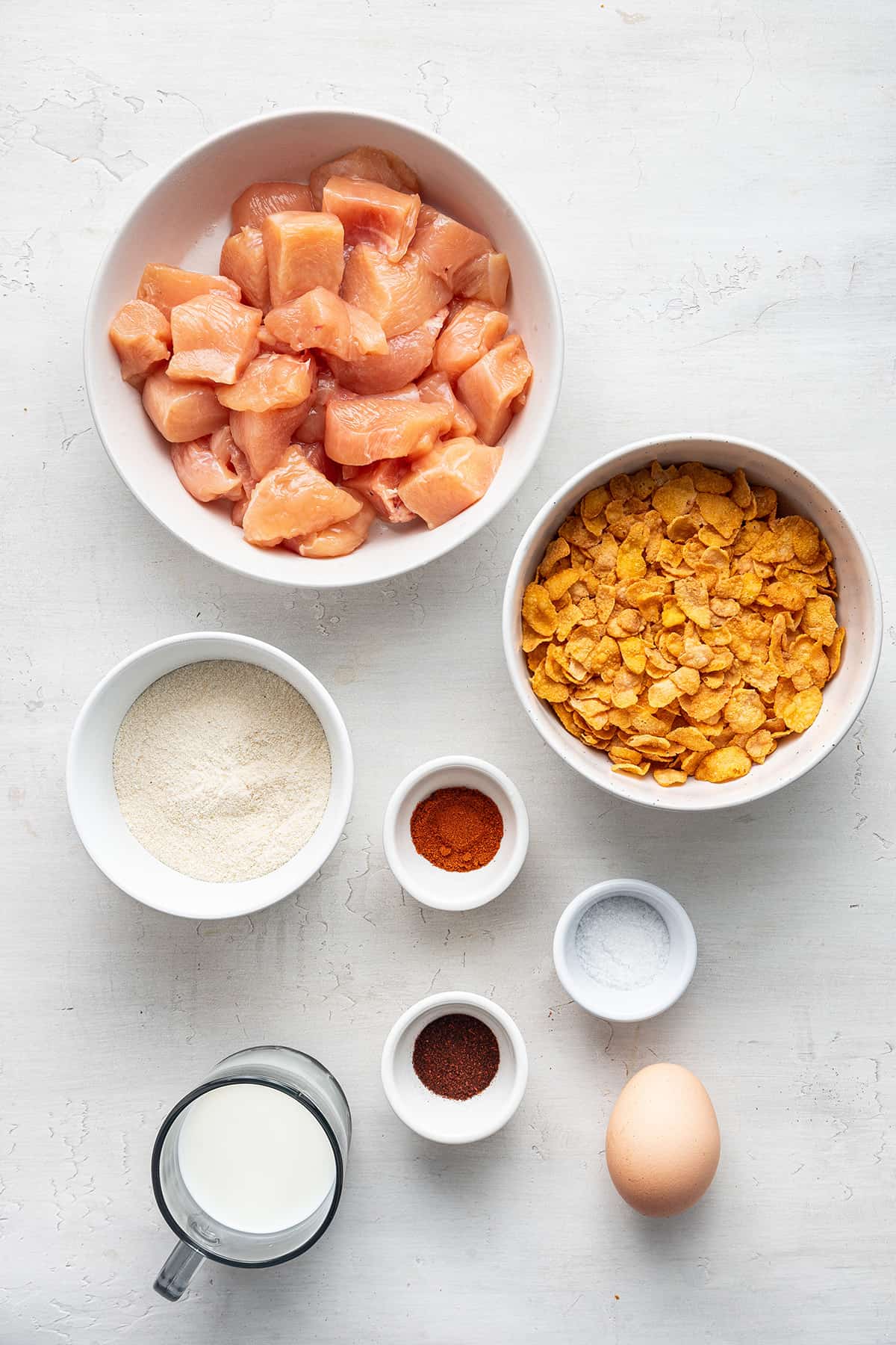 Overhead view of a bowl of uncooked chicken breast chunks, a bowl of corn flakes, a bowl of coconut flour, a bowl of paprika, a bowl of chili powder, a bowl of salt, a pitcher of dairy-free milk, and an egg