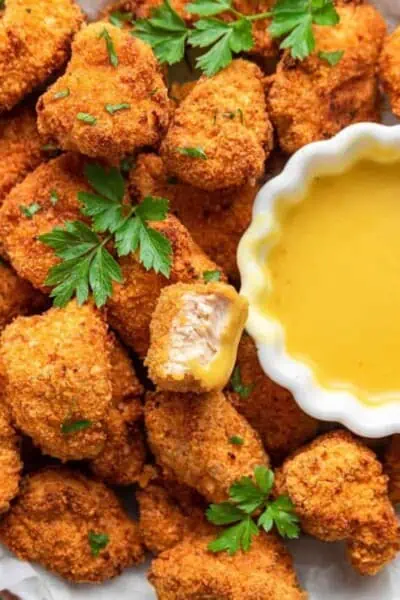 Close up of a pile of popcorn chicken garnished with parsley, with a bite taken out of one piece, and a bowl of dipping sauce