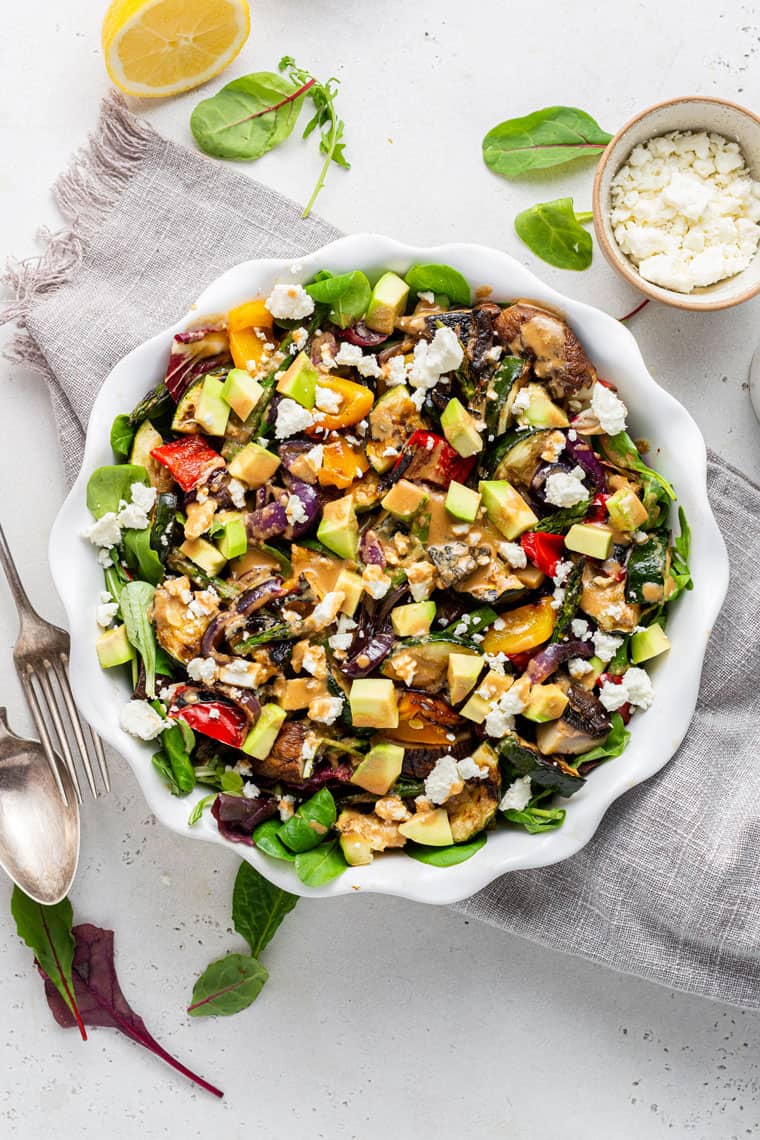 Overhead view of a grilled salad topped with cubes of avocado and crumbled feta, on top of a kitchen towel, next to a few pieces of lettuce, a bowl of feta, a fork, and a spoon. 