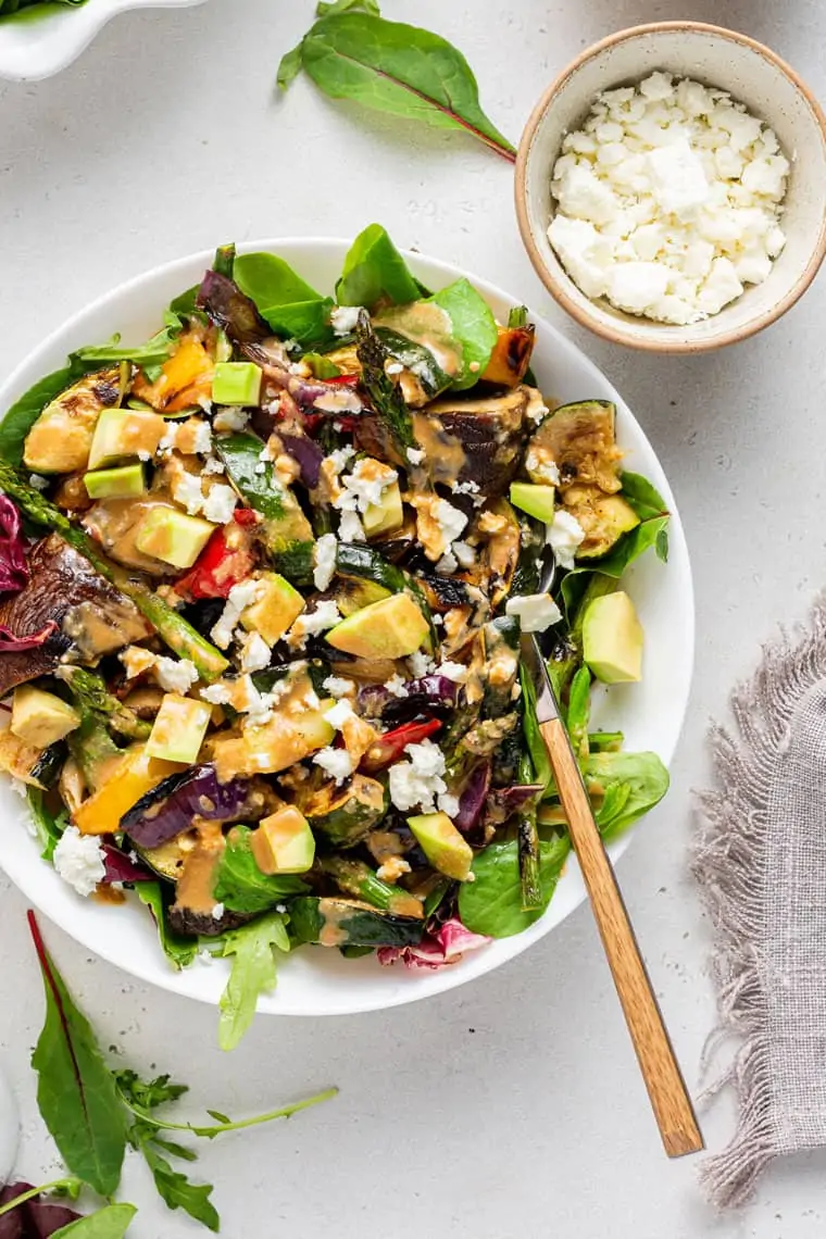 A bowl of grilled salad topped with feta and avocado, with a fork in it, next to a small bowl of crumbled feta.