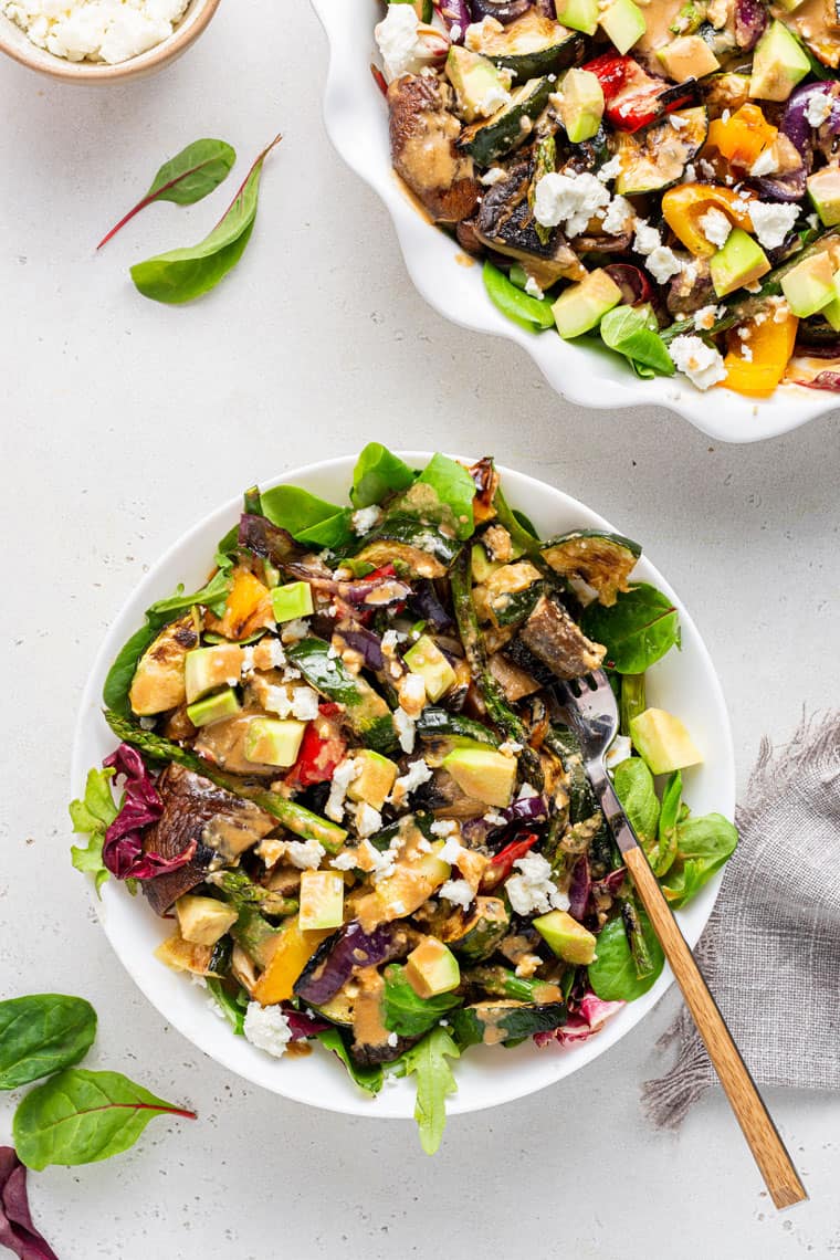 Overhead view of a bowl of grilled salad topped with feta and avocado, with a fork in the bowl, next to some greens and another bowl of salad.