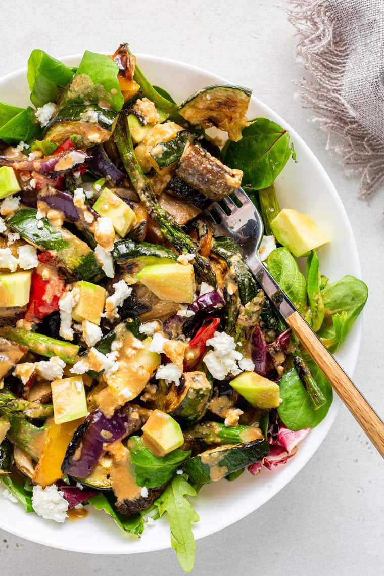 Close up of a fork in a bowl of grilled veggie salad, topped with feta and avocado