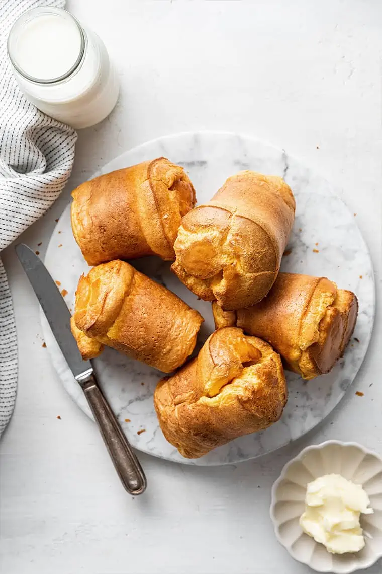 This recipe for gluten-free popovers is moist, buttery, pillowy, and will melt in your mouth, and only takes 45 minutes to make. A plate with five gluten-free popovers on it, with a butter knife on the plate, next to a kitchen towel
