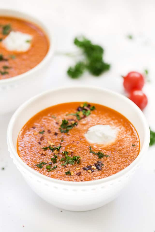 A bowl of roasted red pepper soup topped with chili flakes, parsley, and sour cream, with tomatoes and parsley in the background