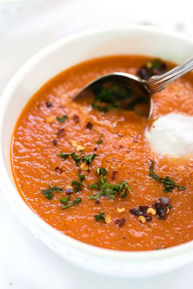 Close up of a bowl of roasted red pepper soup garnished with parsley, sour cream, and chili flakes, with a spoon in it