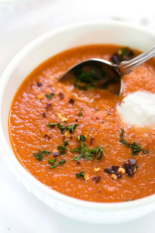 This easy and healthy roasted red pepper soup recipe has fresh tomatoes, quinoa, and lots of spices, and only takes an hour to make. Close up of a bowl of roasted red pepper soup garnished with parsley, sour cream, and chili flakes, with a spoon in it