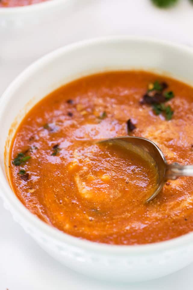 This easy and healthy roasted red pepper soup recipe has fresh tomatoes, quinoa, and lots of spices, and only takes an hour to make. A spoon stirring a bowl of roasted red pepper soup garnished with sour cream, chili flakes, and parsley