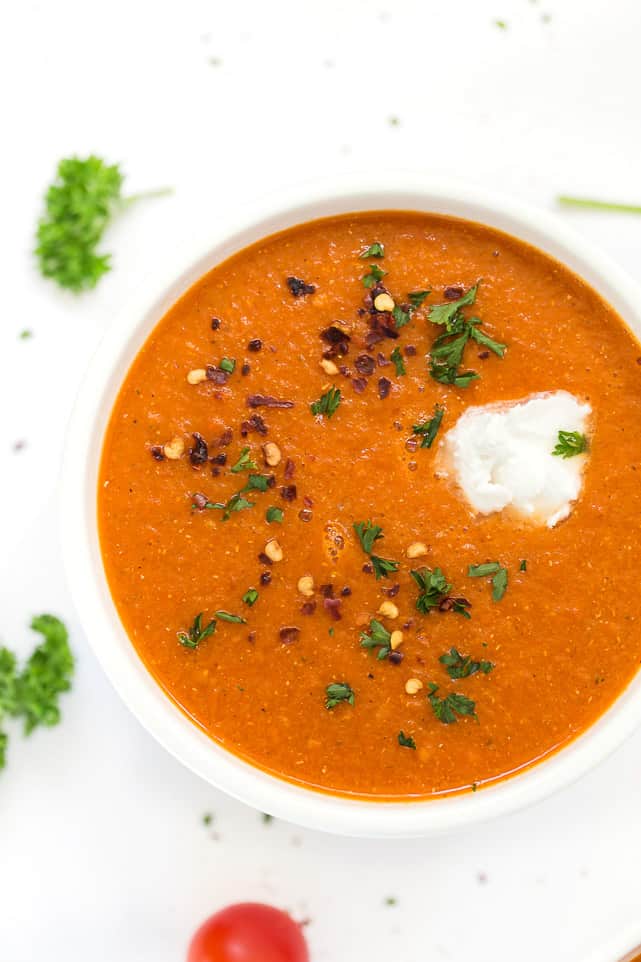 This easy and healthy roasted red pepper soup recipe has fresh tomatoes, quinoa, and lots of spices, and only takes an hour to make. Overhead view of a bowl of roasted red pepper soup topped with parsley, chili flakes, and sour cream, surrounded by parsley