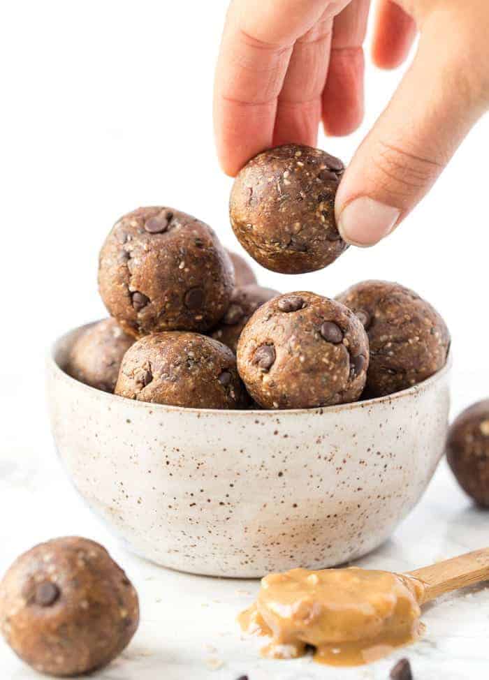 A guide for how to make energy balls, a protein and nutrient-rich snack full of nuts, seeds, and fruit, and easy to take on the go. A hand grabbing a peanut butter chocolate chip ball out of a bowl of them, with a spoonful of peanut butter on the counter