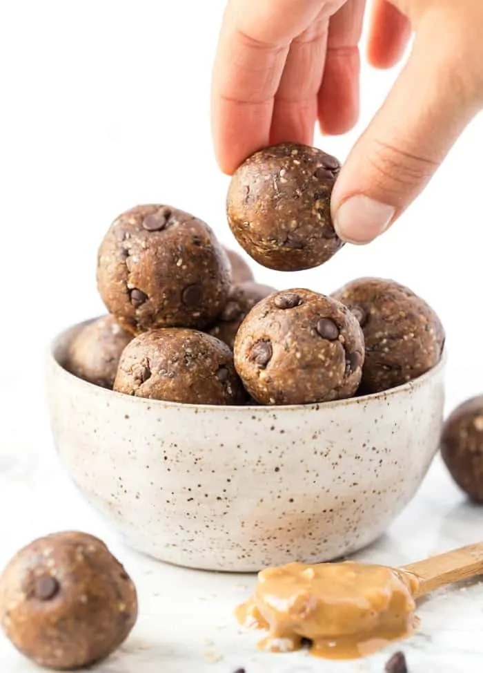 A hand grabbing a peanut butter chocolate chip ball out of a bowl of them, with a spoonful of peanut butter on the counter