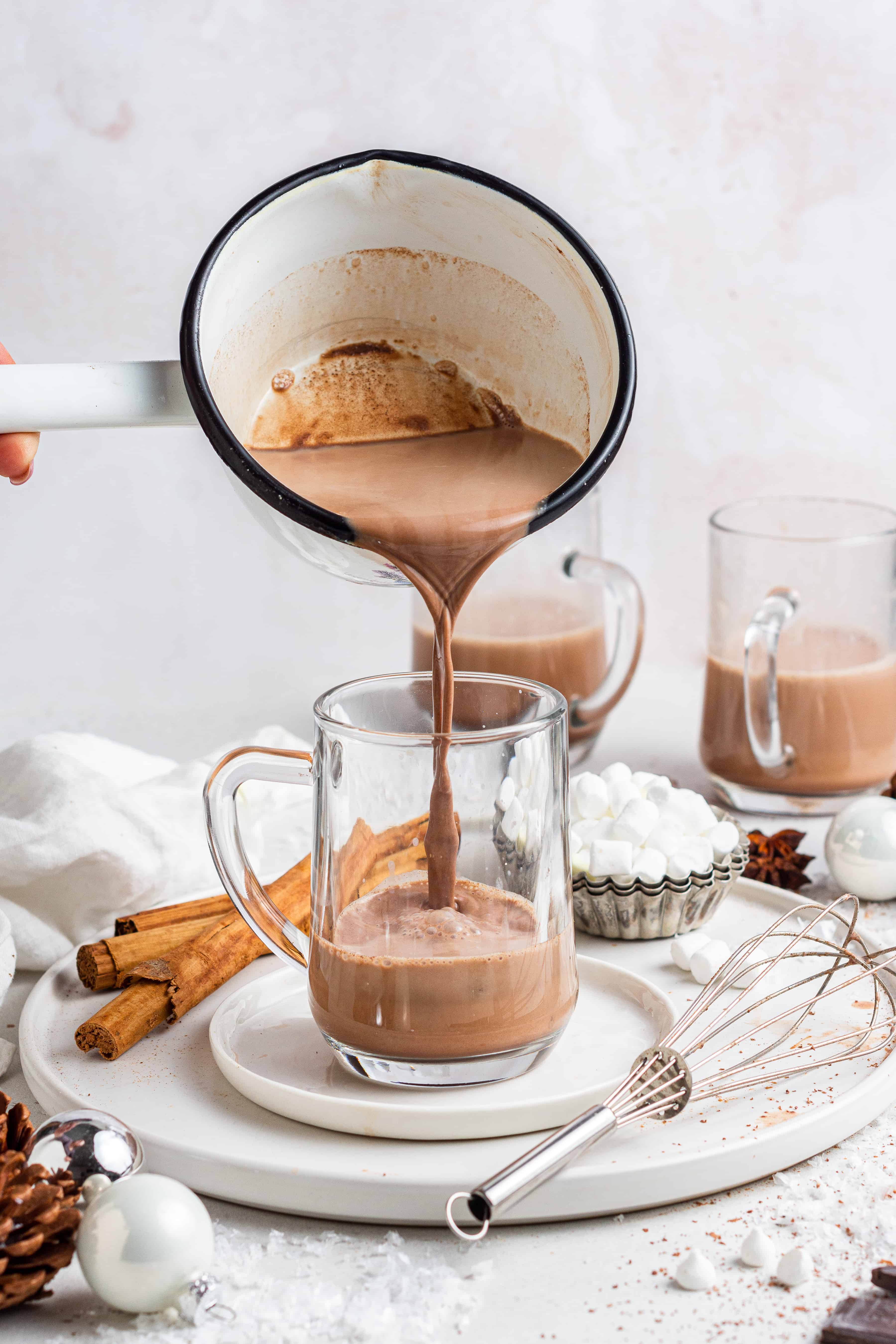 vegan hot chocolate pouring into a glass mug with marshmallows and cinnamon sticks
