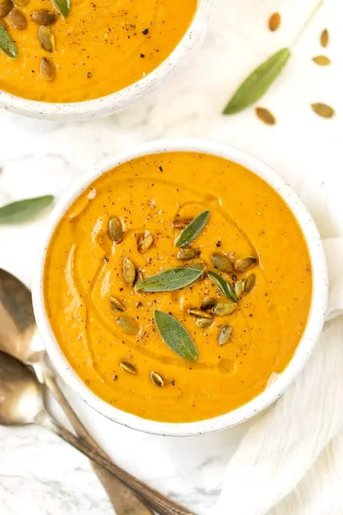 A bowl of roasted pumpkin soup, garnished with pumpkin seeds and sage leaves, on a tablecloth next two two spoons and another bowl of soup