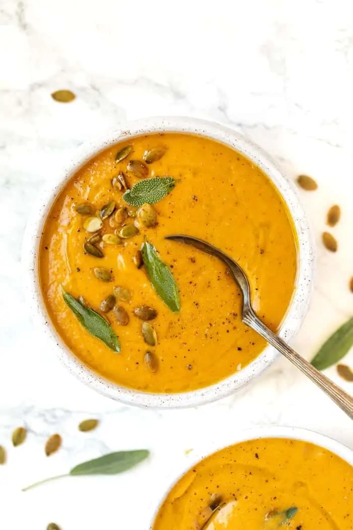 A bowl of roasted pumpkin soup, garnished with safe leaves and pumpkin seeds, with a spoon in it, next to another bowl, with pumpkin seeds and sage leaves scattered around