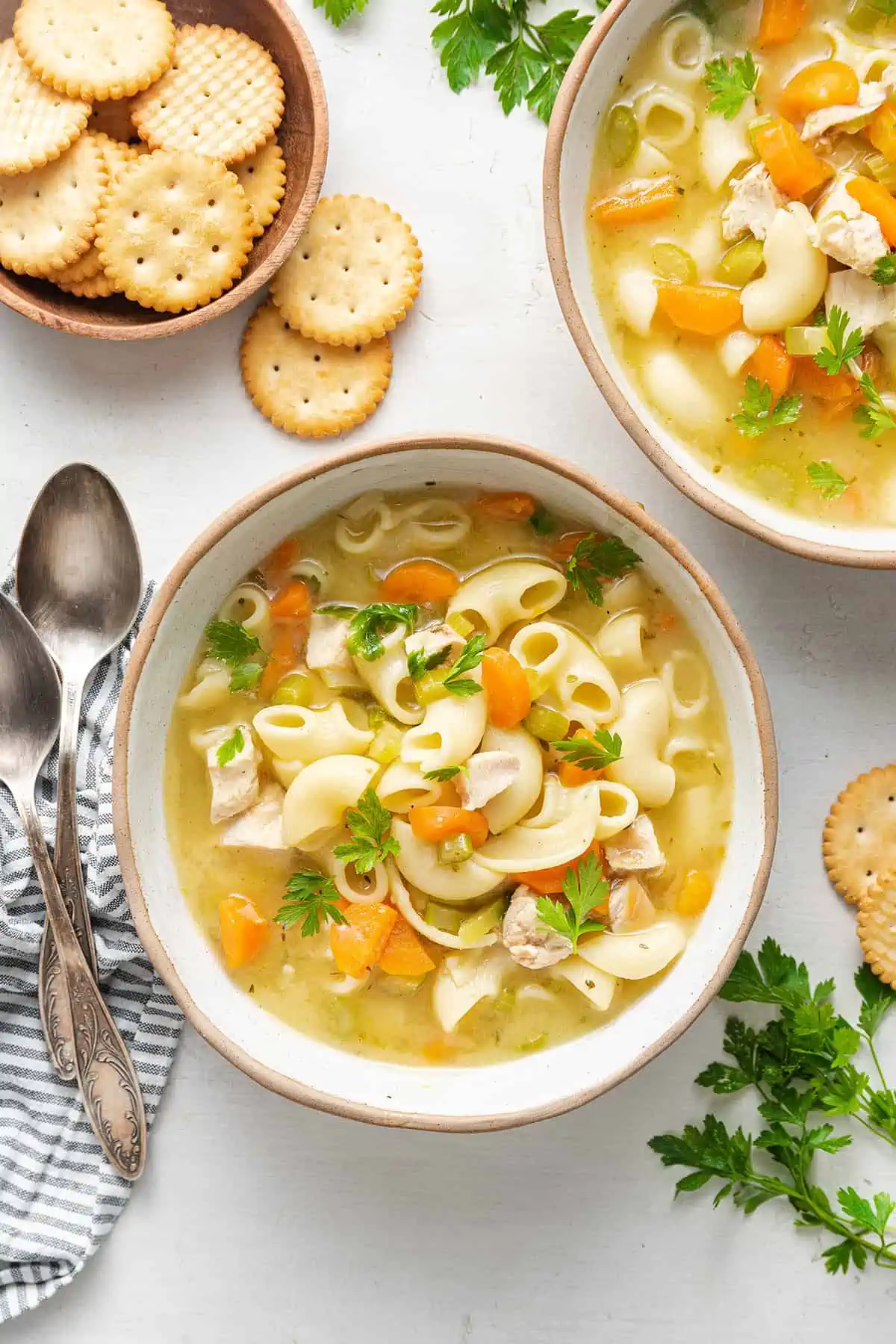 A bowl of chicken noodle soup garnished with parsley, next to another bowl, some round crackers, a napkin, and a spoon