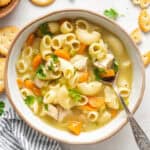 A bowl of chicken noodle soup with a spoon in it, surrounded by a napkin, crackers, and parsley