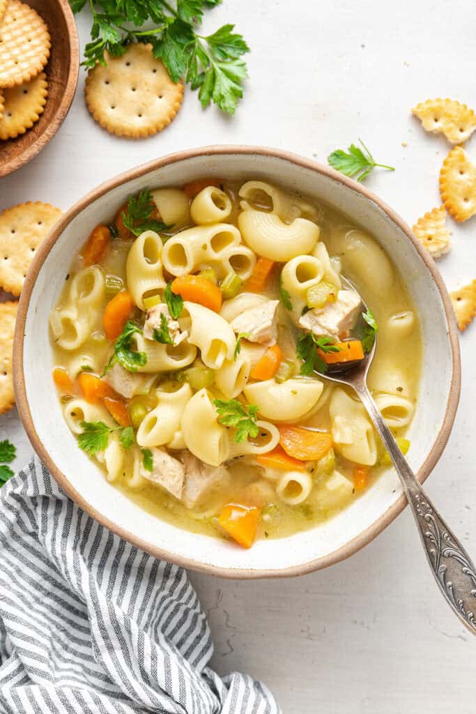 A bowl of chicken noodle soup with a spoon in it, surrounded by a napkin, crackers, and parsley