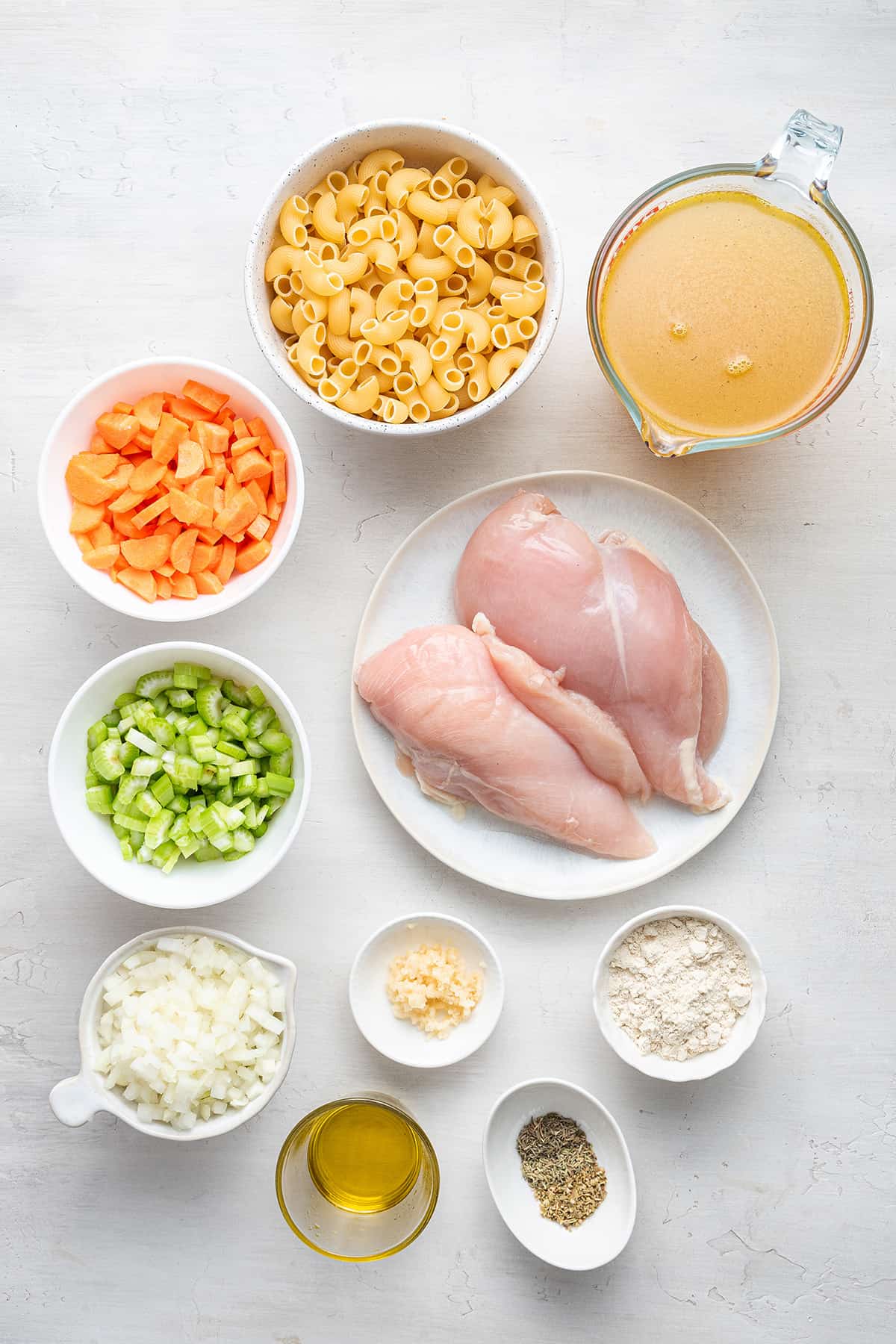 Overhead view of the ingredients needed for chicken noodle soup: chicken breasts, chicken broth, elbow macaroni, carrots, celery, onions, garlic, quinoa flour, spices, and oil