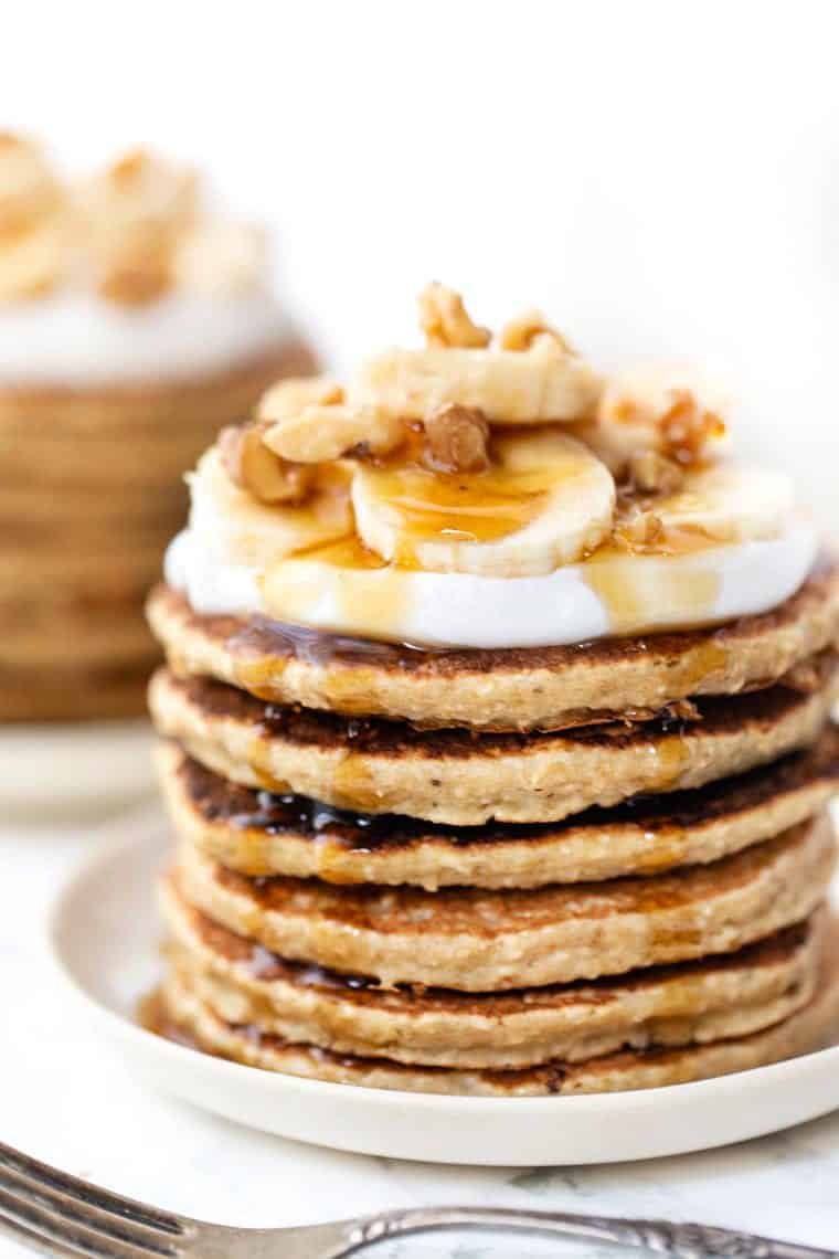 Plate with stack of gluten-free pancakes topped with whipped cream, syrup, sliced banana, and walnuts