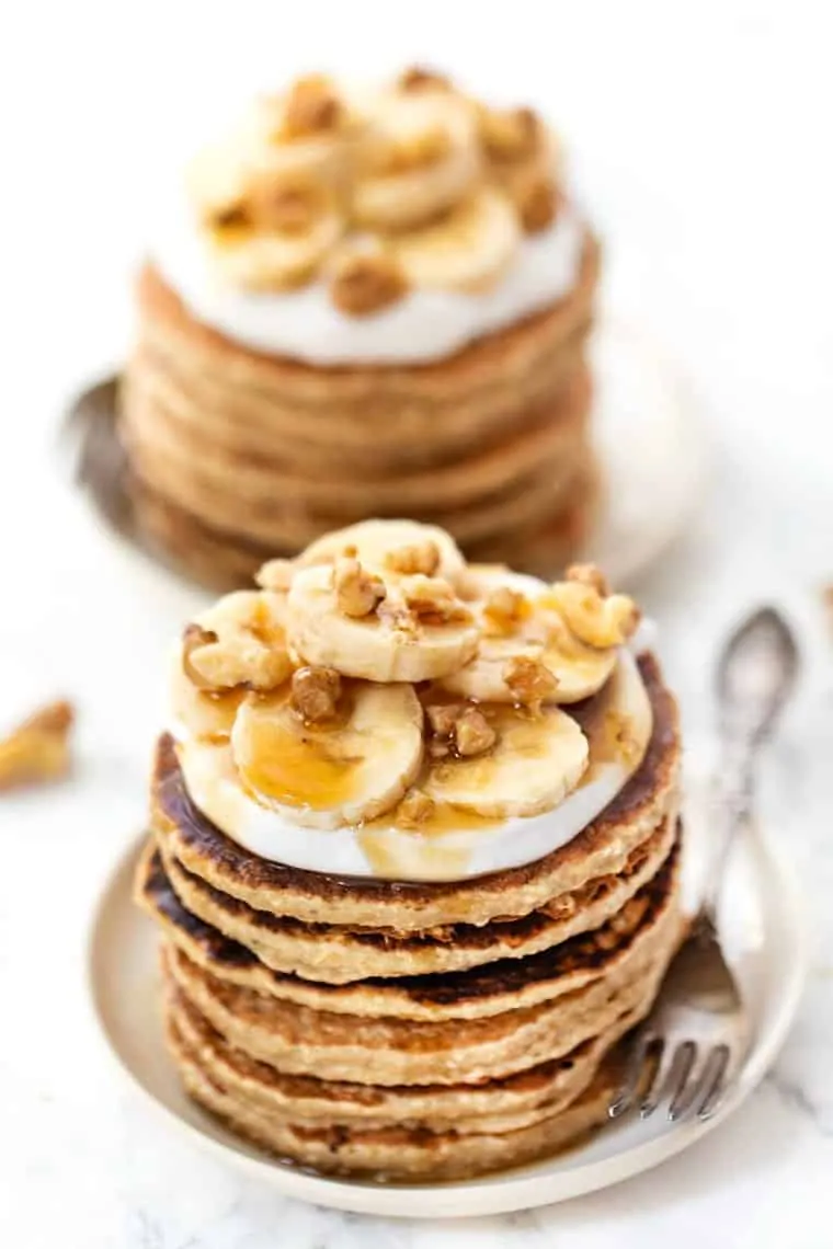Two plates with stacks of quinoa banana pancakes topped with whipped cream, bananas, syrup, and walnuts