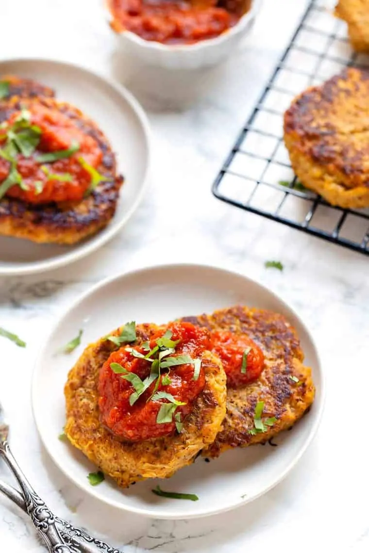 Two spaghetti squash fritters topped with salsa on a white plate, next to another plate of fritters and a bowl of salsa on a white countertop.