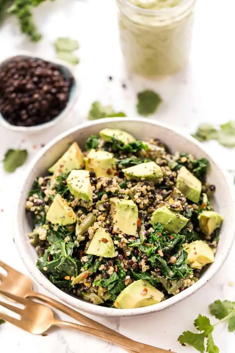 Kale quinoa salad in a bowl next to a fork, with a smaller bowl of black lentils in the background.