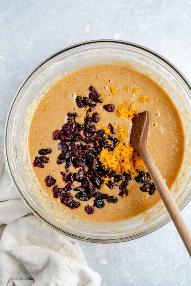 A mixing bowl full of cranberry orange coffee cake, with dried cranberries and orange zest on top, and a wooden spoon in the bowl. 