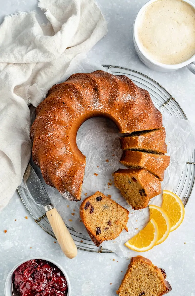 A round coffee cake with a few slices missing and a few slices in place, on a wire rack, next to a cake knife and three slices of oranges