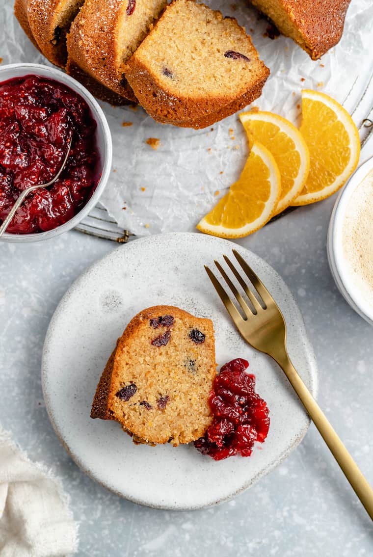 A plate with a slice of cranberry orange coffee cake with cranberry, orange, honey sauce next to it, and a fork on the plate