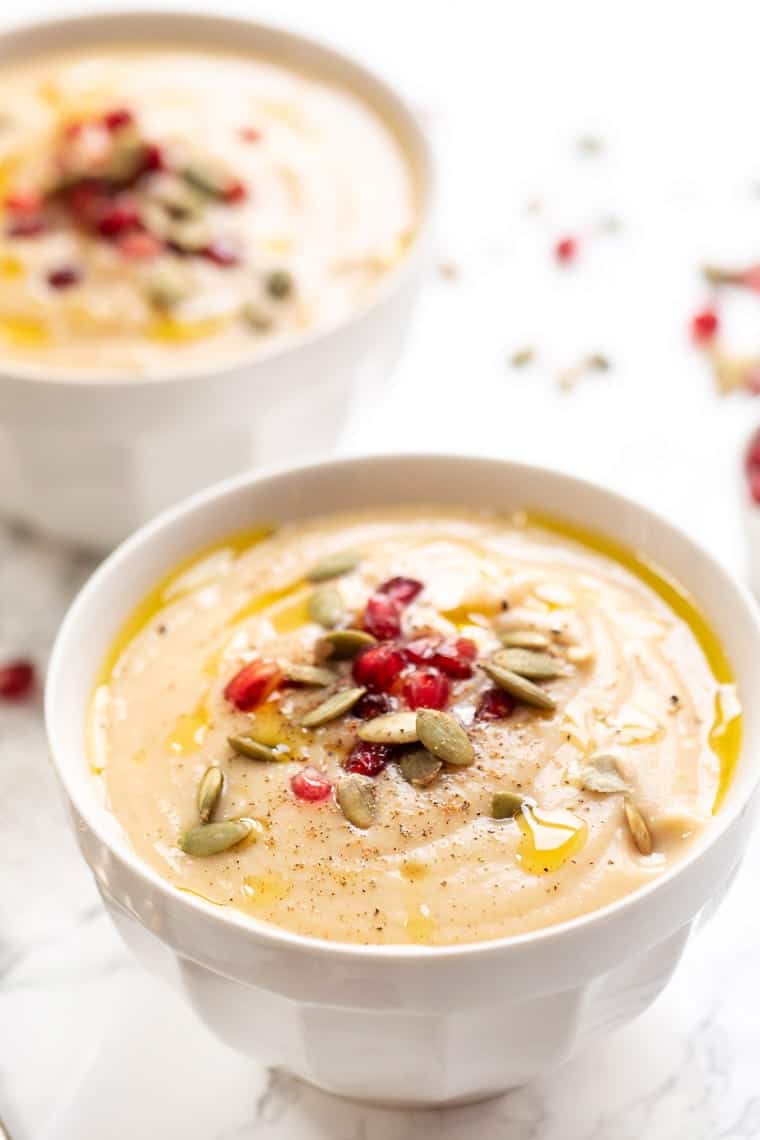A bowl of cauliflower soup, topped with olive oil, pepper, pomegranate seeds, and pumpkin seeds, with another bowl in the background