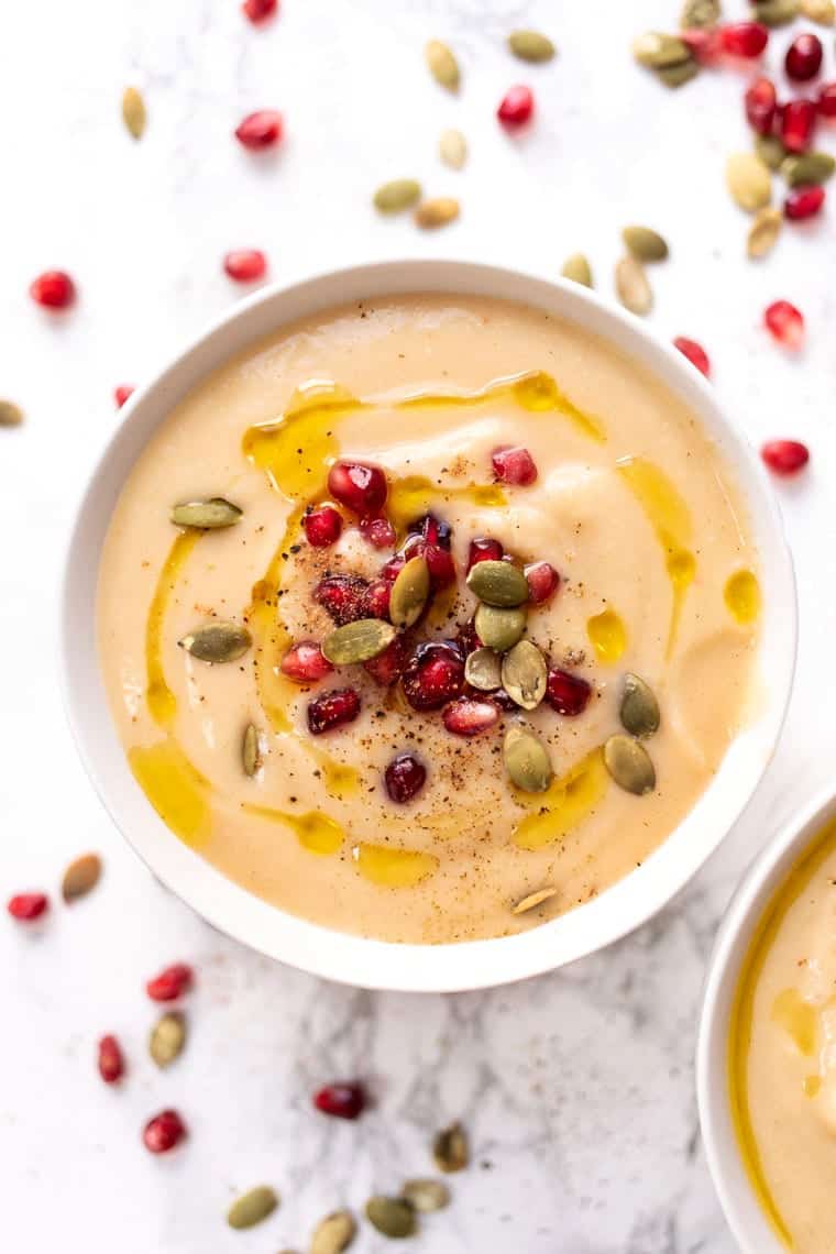 A bowl of caulilflower soup topped with pomegranate seeds, pumpkin seeds, pepper, and olive oil, surrounded by pomegranate seeds and pumpkin seeds
