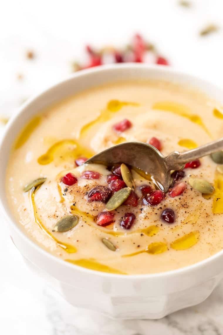 Close up of a bowl of cauliflower soup with a spoon in it, topped with olive oil, pumpkin seeds, pomegranate seeds, and black pepper