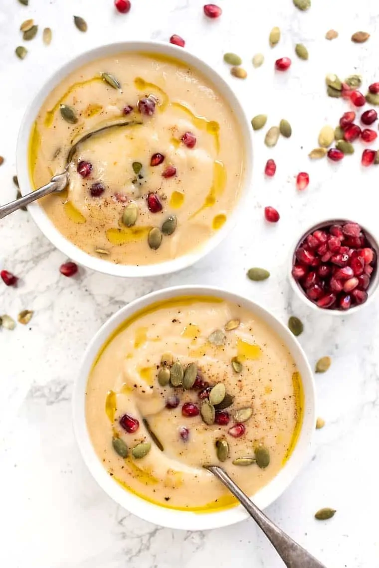 Two bowls of cauliflower soup, topped with pomegranate seeds, pumpkin seeds, and olive oil, with spoons in them, surrounded by pomegranate seeds and pumpkin seeds