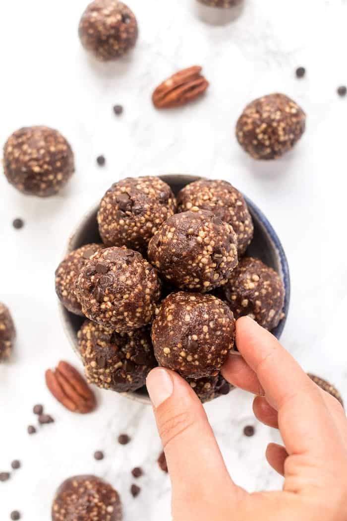 A hand reaching in to grab an energy ball from a bowl of energy balls, with pecans, chocolate chips, and energy balls spilled across the table